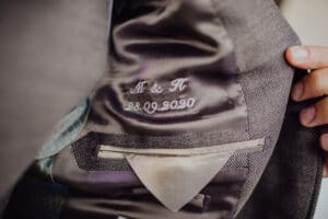 Eine Person hält ein Sakko auf und enthüllt auf dem Innenfutter die aufgestickten Initialen „M & H“ und das Datum „28.09.2020“ – eine subtile Anspielung auf die sorgfältige Hochzeitsplanung. Der Stoff ist dunkel mit einer subtilen Textur und eine Tasche ist sichtbar.