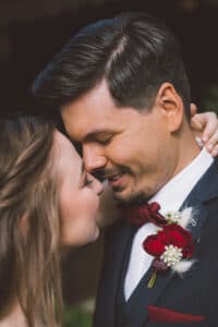 Ein Paar teilt einen intimen Moment, die Gesichter dicht beieinander an ihrem Hochzeitstag. Der Mann trägt einen Anzug mit roter Fliege und Ansteckblume, während die Frau sanft ihre Hand auf seinen Hals legt. In ihren Ausdrücken sind Gefühle der Freude und Liebe deutlich zu erkennen.