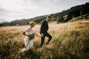 Ein Paar im Hochzeitskleid geht Hand in Hand über eine Wiese und verkörpert damit die Essenz einer perfekten Hochzeit. Die Braut hält einen Blumenstrauß, während der Bräutigam vorangeht. Umrahmt von Bäumen und einem teilweise bewölkten Himmel entsteht eine heitere und romantische Atmosphäre, die ideal für diejenigen ist, die ihren eigenen besonderen Tag planen.
