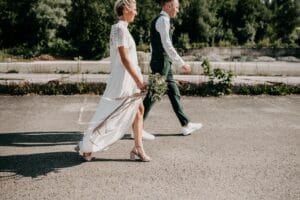 Braut und Bräutigam gehen an einem sonnigen Tag gemeinsam im Freien spazieren. Die Braut in einem weißen Kleid mit einem Blumenstrauß in der Hand und der Bräutigam in einem grünen Anzug genießen ihren Hochzeitsweg auf einer gepflasterten Fläche, umgeben von üppigem Grün.
