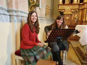 Zwei Frauen sitzen in einer Kirche und sind in Hochzeitsvorbereitungen vertieft. Eine hält eine Gitarre und lächelt von ihrer Bank aus, vor ihr liegen Notenblätter auf einem Ständer. Die andere, die einen roten Mantel und einen grün gemusterten Rock trägt, strahlt, während sie die Einzelheiten der Hochzeit für die bevorstehende Zeremonie besprechen.