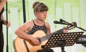 Eine Frau sitzt und spielt Akustikgitarre vor einem Notenständer mit Notenblättern. Vielleicht probt sie für einen besonderen Moment bei einer Hochzeit. Ein Keyboard liegt daneben, während sanftes Tageslicht durch das Fenster fällt.