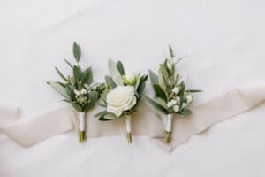 Drei kleine Blumenarrangements mit weißen Rosen und Grünpflanzen, perfekt für eine Hochzeit, sind auf einem hellen Hintergrund ausgelegt. Die Arrangements, ideal für jede Hochzeitsplanung, sind mit weißem Band zusammengebunden und ruhen wunderschön auf einem Streifen hellen Stoffs.