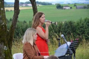 Zwei Frauen treten im Freien bei einer Hochzeit auf. Eine in einem roten Kleid singt ins Mikrofon, während die andere in einer braunen Jacke Keyboard spielt. Sie sind von grünen Feldern und Bäumen umgeben, im Hintergrund sind Hügel und Landschaft zu sehen.