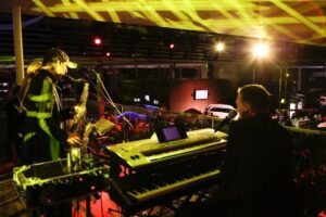 Eine Band, die bei einer Hochzeit auftritt, steht in einem schwach beleuchteten Veranstaltungsort auf der Bühne. Ein Keyboarder und ein Saxophonist sind zu sehen, und bunte Bühnenlichter sorgen für eine energiegeladene Atmosphäre. Im Hintergrund ist das Publikum zu sehen.