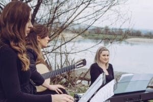 Drei Frauen sitzen im Freien neben einem ruhigen See, umgeben von kahlen Bäumen. Eine hält eine Gitarre, eine andere sitzt vor Notenblättern auf einem Ständer und die dritte schaut zu. Die Umgebung ist friedlich und heiter, als würden sie einen ruhigen Moment der Hochzeitsplanung inmitten der Natur verbringen.