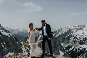 Eine Braut im weißen Kleid hält einen Blumenstrauß auf einem felsigen Bergvorsprung, während ihr Bräutigam im dunklen Anzug ihre Hand hält. Schneebedeckte Gipfel bieten die perfekte Kulisse für ihren Hochzeitsmoment unter dem klaren Himmel.