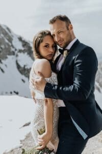 Ein Paar im Hochzeitskleid umarmt sich vor einer Kulisse schneebedeckter Berge und fängt die heitere Atmosphäre ihrer Hochzeit ein. Die Braut in einem Spitzenkleid und mit einem Blumenstrauß in der Hand steht anmutig mit ihrem Bräutigam im dunklen Anzug da, während beide friedlich in die Kamera blicken.