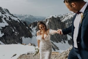 Eine Braut in einem Spitzenbrautkleid lächelt, während sie die Hand ihres Bräutigams im dunklen Anzug hält und ihn unter klarem Himmel durch eine verschneite Berglandschaft führt. Ihre Hochzeit fängt die Essenz von Romantik und Abenteuer in einem atemberaubenden Moment ein.