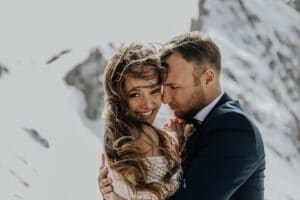 Ein Mann und eine Frau umarmen sich liebevoll auf einem schneebedeckten Berg. Die Frau lächelt und der Kopf des Mannes ruht neben ihrem. Ihr langes Haar weht im Wind, während er einen dunklen Anzug trägt. Vor schneebedeckten Gipfeln und einem klaren Himmel ist es ein malerischer Moment, perfekt für eine unvergessliche Hochzeit.