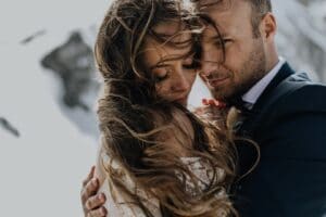 Ein Paar umarmt sich zärtlich im Freien, umgeben von schneebedeckten Bergen. Der Wind zerzaust ihre Haare, während sie in formeller Kleidung von ihrer Hochzeit träumen – der Mann im Anzug und die Frau im weißen Abendkleid.