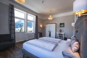 Ein gemütliches Hotelzimmer, ideal für alle, die eine Hochzeit planen, mit einem großen Bett, eleganter Beleuchtung und einem malerischen Fensterblick auf die Berge. In der Ecke stehen ein Kleiderschrank im Vintage-Stil und ein kleiner Schreibtisch mit Spiegel. Das Zimmer hat dunkle Vorhänge und einen Holzboden, perfekt für Hochzeitsvorbereitungen.