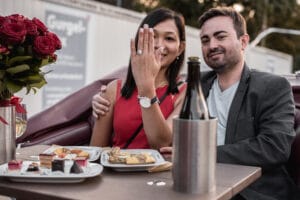 Ein Paar sitzt an einem Tisch mit Tellern voller Essen und einer Flasche Champagner in einem Eiskübel und feiert seine Verlobung. Die Frau in einem roten Kleid zeigt stolz ihren Ring. Ein Strauß roter Rosen schmückt den Tisch, während sie sich freudig umarmen und bereits von ihren Hochzeitsplänen träumen.