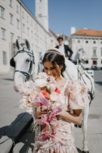 Eine Frau in einem geblümten Kleid hält einen bunten Blumenstrauß und steht vor einem weißen Pferd und einer Kutsche, perfekt für eine elegante Hochzeit. Der Kutscher sitzt auf dem Dach der Kutsche in dieser sonnigen, urbanen Umgebung inmitten historischer Gebäude. Ein malerischer Moment für jede Hochzeitsplanung.
