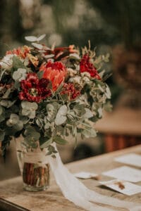 Ein farbenfroher Blumenstrauß in einer Glasvase, perfekt als Hochzeitsdekoration, besteht aus roten und orangefarbenen Blumen, umgeben von üppigen grünen Eukalyptusblättern. Das Arrangement steht auf einem Holztisch neben verstreuten Papieren vor einem unscharfen natürlichen Hintergrund.