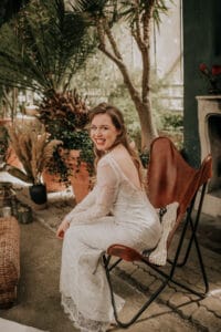 Eine Frau in einem weißen Spitzenkleid sitzt auf einem braunen Stuhl und lächelt inmitten einer üppigen Gartenanlage mit grünen Pflanzen und einfallendem Tageslicht, die die Essenz heiterer Hochzeitsvorbereitungen einfängt.