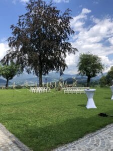Das Hochzeits-Setup im Freien, perfekt für jeden Hochzeits-Enthusiasten, besteht aus weißen Stühlen in Reihen auf einer Wiese. Ein großer Baum und kleinere Bäume rahmen die Szene ein, mit Bergen und einem teilweise bewölkten Himmel als Hintergrund. Im Vordergrund steht ein bezaubernder weißer Tisch.