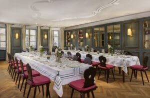 Ein langer Esstisch ist mit weißen Tischdecken, Glaswaren und Blumen in einem Raum gedeckt, der sich ideal für Hochzeitsfeiern eignet. Die Stühle haben rote Sitzflächen und die Wände sind mit Regalen, Spiegeln und Lampen geschmückt. Große Fenster lassen natürliches Licht in den eleganten Raum.