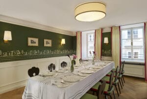 Ein gut dekorierter Speisesaal, der sich perfekt für eine Hochzeitsfeier eignet, verfügt über einen langen Tisch mit weißer Tischwäsche, elegantem Glasgeschirr und Tellern. Die olivgrünen Wände ergänzen gerahmte Kunst und eine große Deckenleuchte. Zwei Fenster mit Vorhängen füllen den Raum mit natürlichem Licht, ideal für die Hochzeitsplanung.