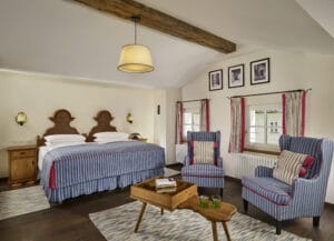 Ein gemütliches Schlafzimmer, perfekt für einen Hochzeitsplanungs-Rückzugsort, mit zwei gestreiften Sesseln, einem hölzernen Couchtisch und zwei Einzelbetten mit gestreifter Tagesdecke. Holzbalken, gerahmte Fotos und große Fenster mit gestreiften Vorhängen verleihen dem Raum Charme. Von der Decke hängt eine warme Lampe.