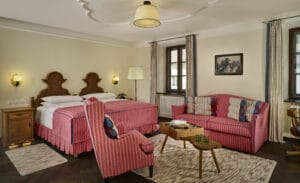 Ein gemütliches Hotelzimmer, perfekt für einen Hochzeitsausflug, mit zwei Betten mit rot-weiß gestreifter Bettwäsche. Es gibt ein passendes Sofa und einen Sessel, einen hölzernen Couchtisch und kleine, von Lampen beleuchtete Nachttische. Große Fenster mit Vorhängen erhellen das warme Vintage-Dekor, ideal für romantische Momente.
