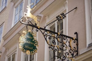 Ein kunstvolles Schild aus Schmiedeeisen und Gold für „Hotel Goldener Hirsch“ hängt an einem Gebäude mit großen Fenstern. Eine goldene Hirschfigur sitzt oben auf dem Schild und verleiht ihm eine klassische, elegante Note – ein einladender Anblick für alle, die eine Hochzeitsfeier oder eine sorgfältige Hochzeitsplanung planen.