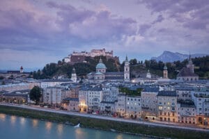 Eine malerische Aussicht auf Salzburg, Österreich, perfekt als Hochzeitskulisse, zeigt die historische Festung Hohensalzburg auf einem Hügel. Im Vordergrund fließt die Salzach, mit barocker Architektur und Kirchtürmen unter einem bewölkten Himmel und Bergen in der Ferne.