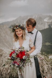 Ein Paar posiert auf einem Berg mit malerischer Kulisse und feiert seine Hochzeit. Die Frau in Spitzenkleid und Blumenkrone hält einen farbenfrohen Blumenstrauß. Der Mann in Hosenträgern und Fliege steht hinter ihr. Beide wirken fröhlich und entspannt inmitten der Natur.