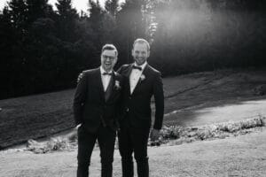 Zwei Männer in Anzügen stehen lächelnd und mit den Armen umeinander auf einer grasbewachsenen Außenfläche und fangen die Freude ihres Hochzeitstages ein. Einer trägt eine Brille und beide sind in formeller Kleidung. Sonnenlicht fällt durch die Bäume und verstärkt die freudige und feierliche Stimmung dieses besonderen Moments der Hochzeit.