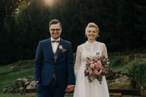 Ein Paar, bereit für seine Hochzeit, steht draußen. Der Bräutigam trägt einen marineblauen Anzug mit Fliege und die Braut ein weißes Spitzenkleid mit rosa Blumen in der Hand. Sie lächeln glücklich zwischen Bäumen und Felsen und fangen den perfekten Moment ihrer Hochzeitsreise ein.