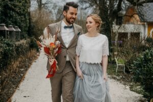 Ein Paar, das seine Hochzeit feiert, geht lächelnd im Freien spazieren. Der Mann trägt einen beigen Anzug und hält einen rot verpackten Blumenstrauß in der Hand. Die Frau trägt ein weißes Oberteil und einen grauen Rock und hält ihren Rock fest, während sie einen von Grünpflanzen gesäumten Kiesweg entlangschlendern.