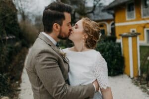 Eine Braut in einem weißen Spitzenkleid und ein Bräutigam in einem braunen Anzug küssen sich auf einem Kiesweg. Im Hintergrund sieht man Grün und ein gelbes Haus mit weißen Verzierungen. Die ruhige Szene fängt die romantische Essenz ihrer Hochzeit ein und schafft unvergessliche Erinnerungen an ihren besonderen Tag.