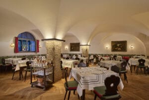 Ein gemütliches, elegant dekoriertes Restaurant-Interieur, perfekt für Hochzeitsplanung, mit weißen Tischdecken und dunklen Holzstühlen. Warmes Licht hebt die Steinsäulen und die gewölbte Decke hervor, während ein Fenster mit roten Vorhängen und gerahmten Gemälden die Wände schmückt.