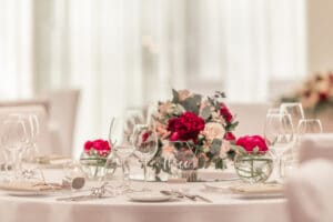 Elegante Tischdekoration mit leeren Weingläsern und Tellern auf einer weißen Tischdecke. Perfekt für eine Hochzeit, zeigt das Mittelstück ein Blumenarrangement mit roten und weißen Blumen, akzentuiert durch Grünpflanzen. Der sanft verschwommene Hintergrund verstärkt die ruhige Atmosphäre, ideal für Ihre Hochzeitsplanung.
