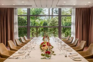 Ein langer, eleganter Esstisch, der für eine exquisite Hochzeit gedeckt ist, besteht aus Weingläsern, weißen Tellern und Besteck. Rote und weiße Blumenarrangements mit Kerzen zieren die Mitte. Große Fenster geben den Blick auf üppiges Grün frei, während Samtstühle auf beiden Seiten stehen – eine perfekte Kulisse zum Heiraten.