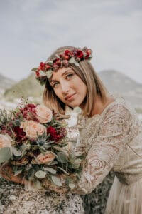 Eine Braut in einem Spitzenhochzeitskleid und mit einer Blumenkrone blickt in die Kamera und verkörpert eine perfekte Hochzeitsvision. Sie hält einen Strauß pfirsichfarbener und burgunderfarbener Blumen in der Hand, umrahmt von Bergen und einem klaren Himmel, und fängt einen eleganten Moment inmitten ihrer Hochzeitsplanung ein.