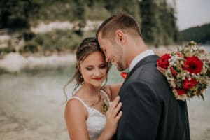 An einem ruhigen See, in einem zärtlichen Moment umarmt, feiert das Paar seine Hochzeit. Die Frau lächelt sanft und hält rote und weiße Blumen in der Hand, während beide formelle Kleidung tragen. Die Stirn des Mannes ruht sanft an ihrer, und sie genießen dieses friedliche Zwischenspiel zwischen Bäumen – eine Szene aus ihrer sorgfältig ausgearbeiteten Hochzeitsplanung.