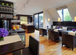 Moderne Frühstückslounge mit Holzboden und Tischen mit braunen Stühlen, perfekt für einen Hochzeitsplanungsbrunch. Frische Säfte, Gebäck auf der Theke und lila Blumen verleihen einen Hauch von Eleganz. Große Glasfenster durchfluten den Raum mit natürlichem Licht.
