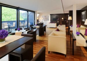 Moderne Lounge mit Holzboden, cremefarbenen Sofas und dunkelbraunen Stühlen, mit violetten Blumenarrangements auf den Tischen. Große Fenster geben den Blick ins Grüne frei, wie geschaffen für Ihre Hochzeitsträume. Wände in neutralen Tönen und minimalistisches Dekor schaffen eine elegante Atmosphäre, die perfekt für die Hochzeitsplanung ist.