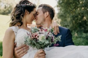 Braut und Bräutigam umarmen sich innig und schauen sich inmitten üppigen Grüns in die Augen. Die Braut hält einen Blumenstrauß in der Hand, der die Essenz des Heiratens verkörpert. Umgeben von der Schönheit der Natur teilt das Paar einen freudigen, romantischen Moment, der ihren Hochzeitstag unvergesslich macht.