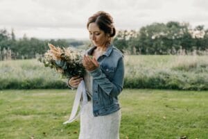 Eine Frau in Brautkleid und Jeansjacke steht auf einer Wiese und hält einen Strauß rustikaler Blumen in der Hand. Die Szene strahlt Hochzeitszauber aus, und der von Bäumen gesäumte Horizont unter einem bewölkten Himmel verleiht diesem spontanen Heiratsmoment noch mehr Charme.