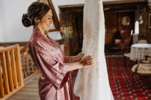 Eine Frau in einem rosa Morgenmantel lächelt, während sie ein weißes Brautkleid in den Händen hält und verkörpert damit die Freude an der Hochzeitsplanung. Sie steht in einem warm beleuchteten, rustikalen Raum mit Holzbalken, Vintage-Möbeln und einem rot gemusterten Teppich.