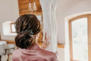 Eine Frau in einem rosa Gewand mit einem dekorativen Haarteil steht drinnen und betrachtet ein hängendes Spitzenhochzeitskleid. Der Raum, perfekt für eine Hochzeit, hat Holzelemente und gewölbte Fenster, durch die Sonnenlicht hereinströmt, während sie von ihrer bevorstehenden Hochzeit träumt.