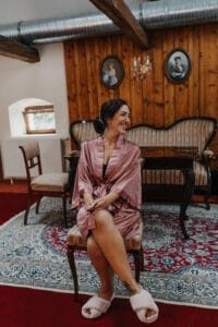 Eine Frau in einem rosa Bademantel und flauschigen Pantoffeln sitzt lächelnd auf einem Stuhl in einem gemütlichen Raum mit Holzwänden, Vintage-Porträts und antiken Möbeln und träumt vielleicht von ihrer bevorstehenden Hochzeit. Ein gemusterter Teppich bedeckt den roten Teppichboden und verleiht dem Raum zusätzlichen Charme.