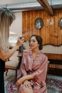 Eine Frau sitzt in einem Satinmantel auf einem Stuhl und bereitet sich auf ihre Hochzeit vor, während eine andere Frau ihr Haar mit Haarspray frisiert. Die Holzwände des Raums, die Vintage-Möbel und die Porträts in ovalen Rahmen schaffen eine rustikale und elegante Atmosphäre, die perfekt für einen solchen Anlass ist.