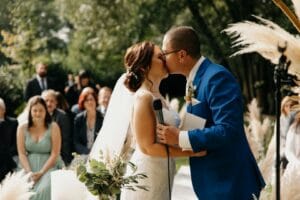 Bei einer malerischen Hochzeit im Freien küssen sich Braut und Bräutigam zärtlich. Der Bräutigam in einem schicken blauen Anzug hält ein Mikrofon, während die Braut in ihrem weißen Kleid mit dem Blumenstrauß in der Hand glänzt. Sitzende Gäste schauen inmitten üppigen Grüns und Pampasgrass zu und feiern diesen wunderschönen Tag der Hochzeit.