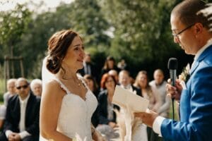 Bei einer wunderschönen Hochzeit im Freien gibt sich ein Paar in der Sonne das Ja-Wort. Die Braut in ihrem weißen Kleid blickt den Bräutigam in seinem blauen Anzug liebevoll an, während er das Mikrofon umklammert. Die Gäste beobachten von ihren Plätzen aus einen perfekten Moment, der durch eine sorgfältige Hochzeitsplanung geprägt wurde.