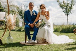 Eine Braut in einem weißen Kleid und Schleier sitzt neben einem Bräutigam im blauen Anzug auf einem Holzstuhl im Freien und wischt sich während ihrer Hochzeit mit der Hand Tränen ab. Das emotionale Paar ist von Gras und Bäumen umgeben und wunderschön eingefangen, während sie diesen Moment der Liebe und Hingabe genießen.