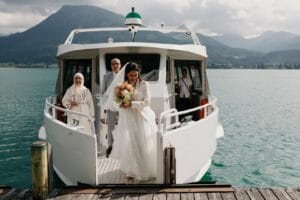 Eine Braut in einem weißen Kleid steigt mit einem Blumenstrauß in der Hand von einem Boot auf einen Holzsteg. Mit Hochzeitsträumen in den Augen stehen zwei Menschen in traditioneller Kleidung und ein Besatzungsmitglied hinter ihr. Die malerische Kulisse zeigt Berge und einen See unter einem bewölkten Himmel.