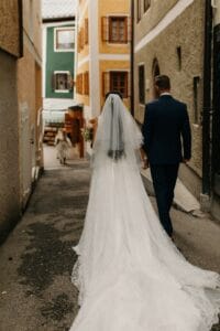 Eine Braut in einem langen weißen Brautkleid und Schleier geht neben einem Mann im Anzug eine schmale Straße entlang. Ihnen folgt ein Kind. Die farbenfrohen Gebäude mit hölzernen Fensterläden tragen zur malerischen Hochzeitskulisse bei, perfekt für alle, die von Hochzeiten träumen.